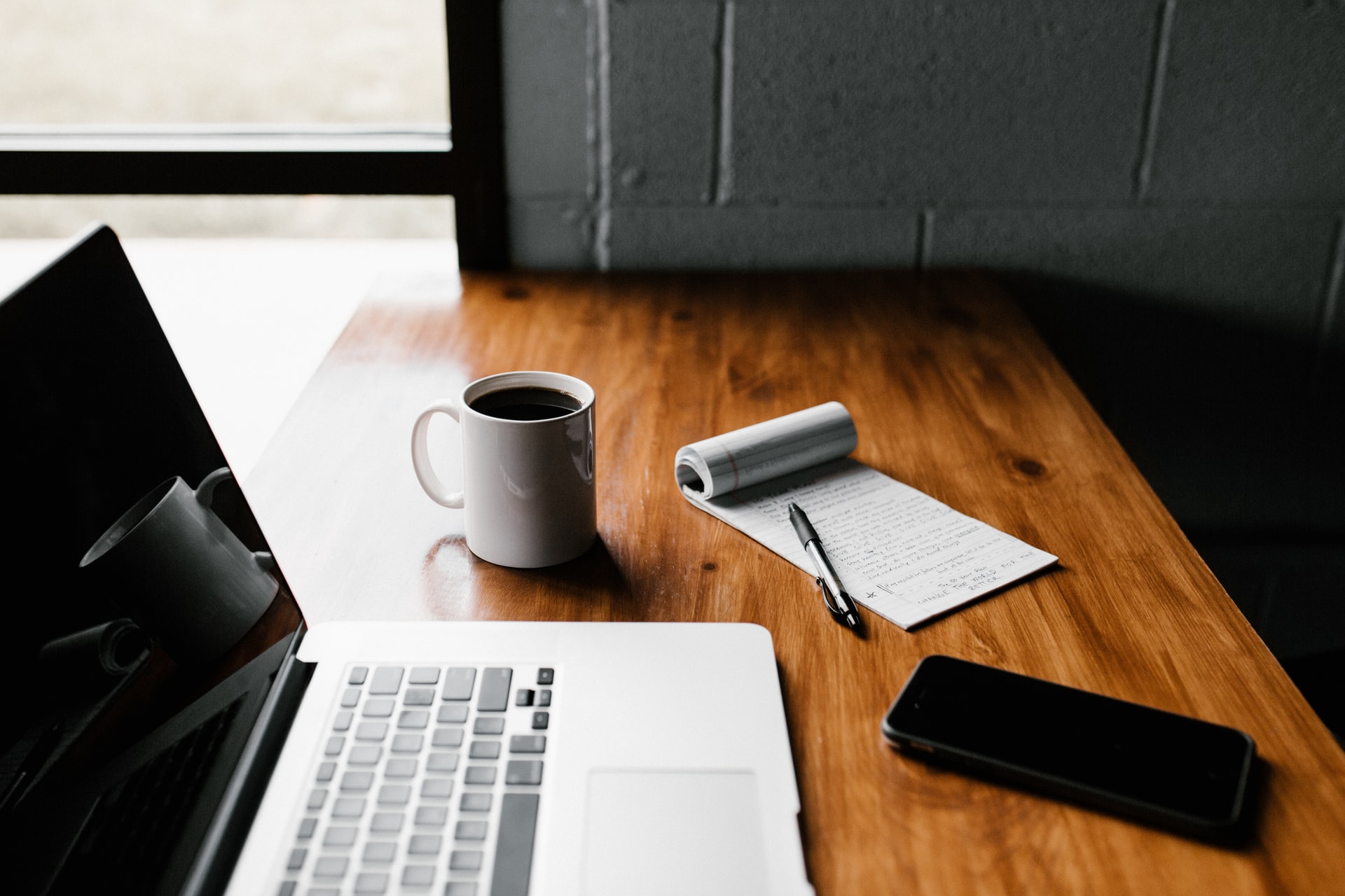 An open laptop sits on a wooden desk nest to a mug of coffee and an open notebook