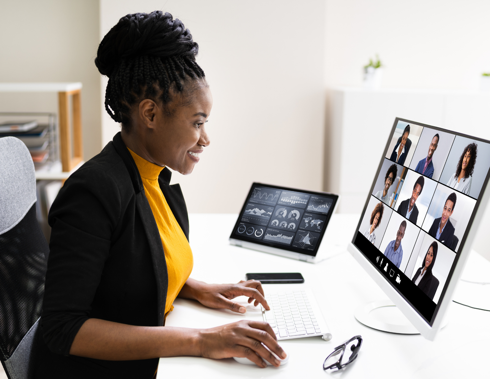 A Black woman wearing a yellow shirt and a black blazer sits in fron of her computer screen talking to several faces in boxes