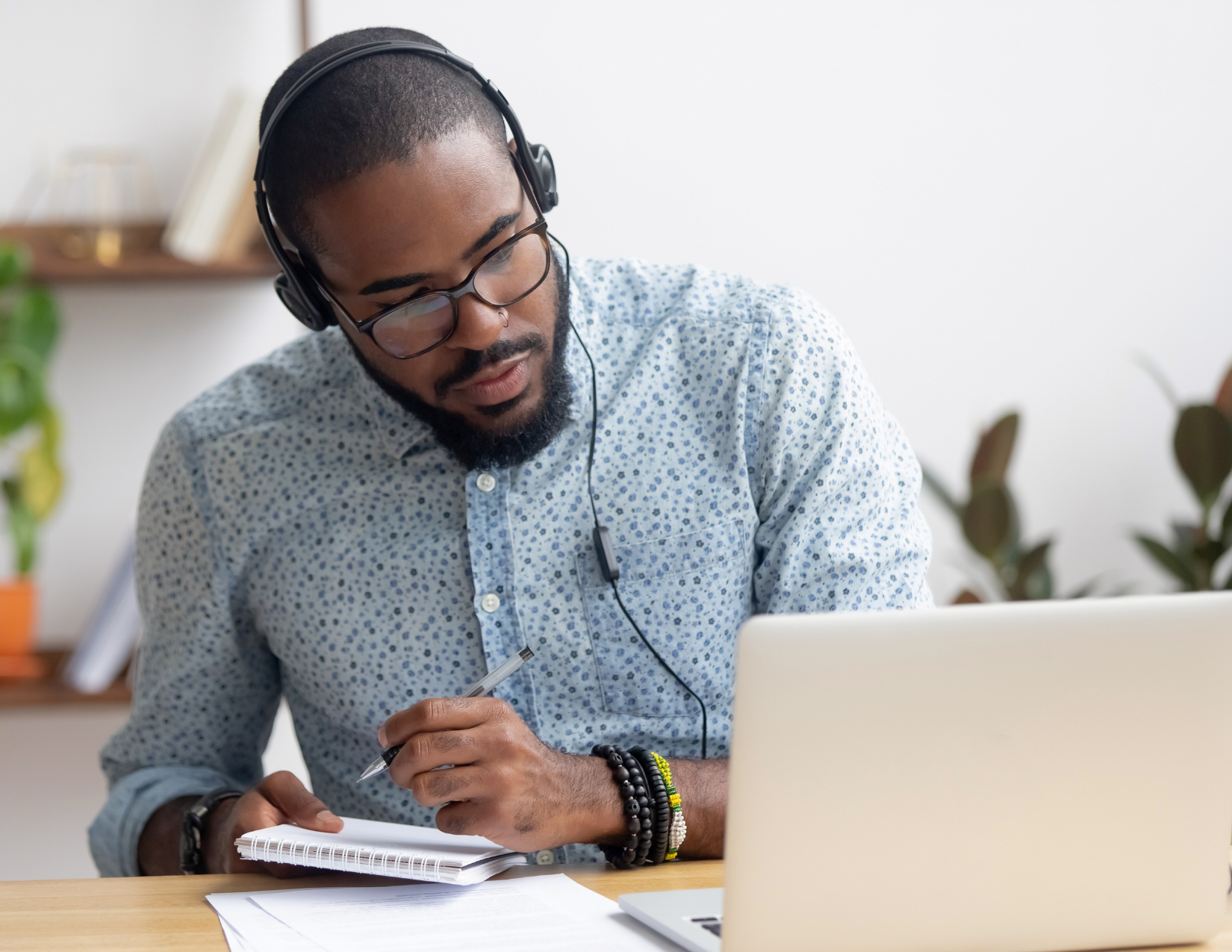 A Black man in a blue button up dress shirt sits in front of a laptop taking notes