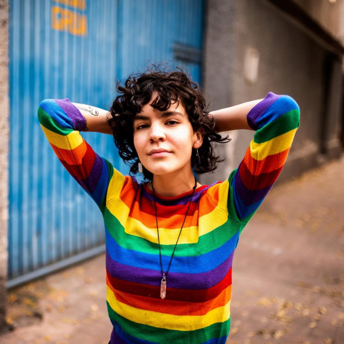 A woman stands with her hands folded behind her head wearing a rainbow striped sweater